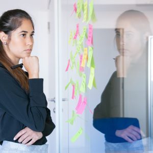 Thoughtful Latin businesswoman holding marker and reading notes on glass wall. Focused confident pretty female worker in suit thinking about idea for project. Brainstorming and business concept