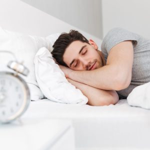 Photo of handsome man having stubble and wearing casual clothes sleeping at home in bed with clock on nightstand
