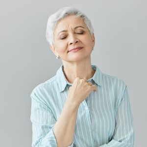 Studio image of beautiful gray haired woman pensioner in blue striped shirt closing eyes and smiling peacefully, enjoying good classical music, having nostalgic facial expression, holding at her chest
