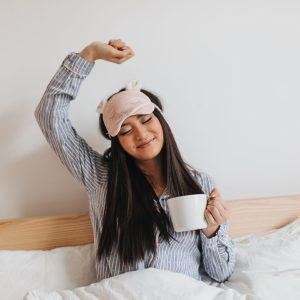 Long-haired woman in blue pajamas streches sweetly after sleeping. Girl holding cup